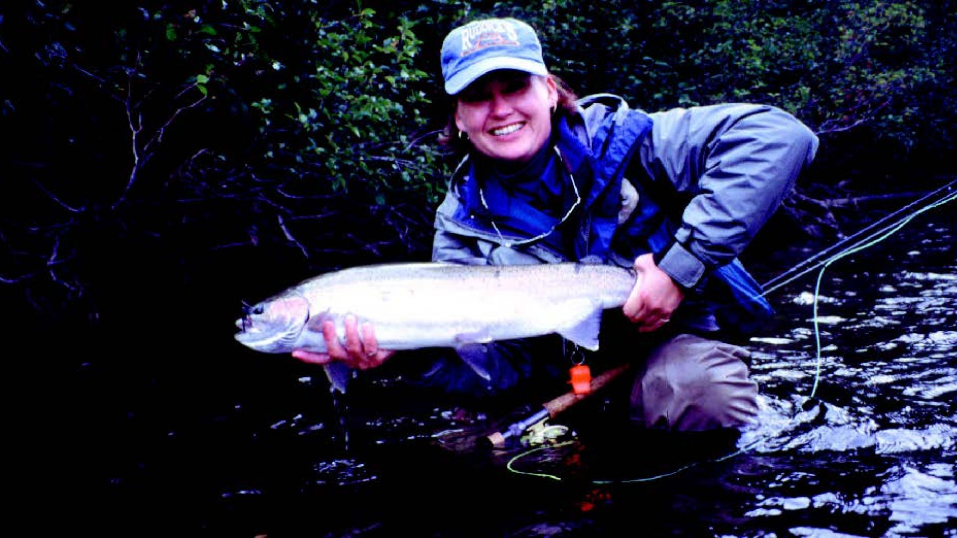 Kathy Ruddick crouches down in her waders to pose with a large fish she's just caught.